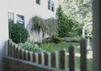 view of a garden in the backyard with white fence in the forefront and bird bath and arbor in the background
