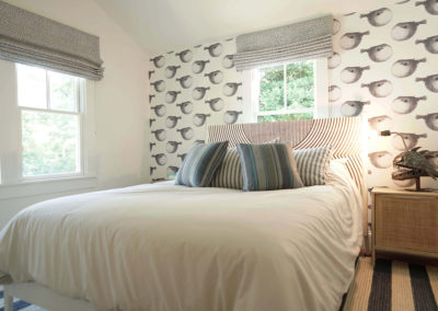 bedroom with queen size bed with white comforter and striped throw pillows and striped headboard, nightstand to the right with large fish structure as the lamp, and matching graphic wallpaper on the back wall