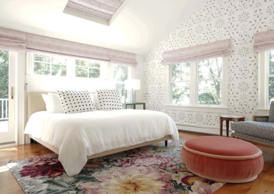 master bedroom with graphic wallpaper, pink window shades, large floral rug, white king size bed, and peach colored round couch with blue chair in right corner